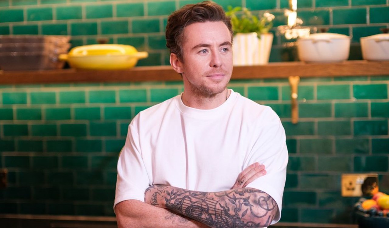 Man (Danny Jones) wearing a white t-shirt, arms folded, stood in front of green brick tiles